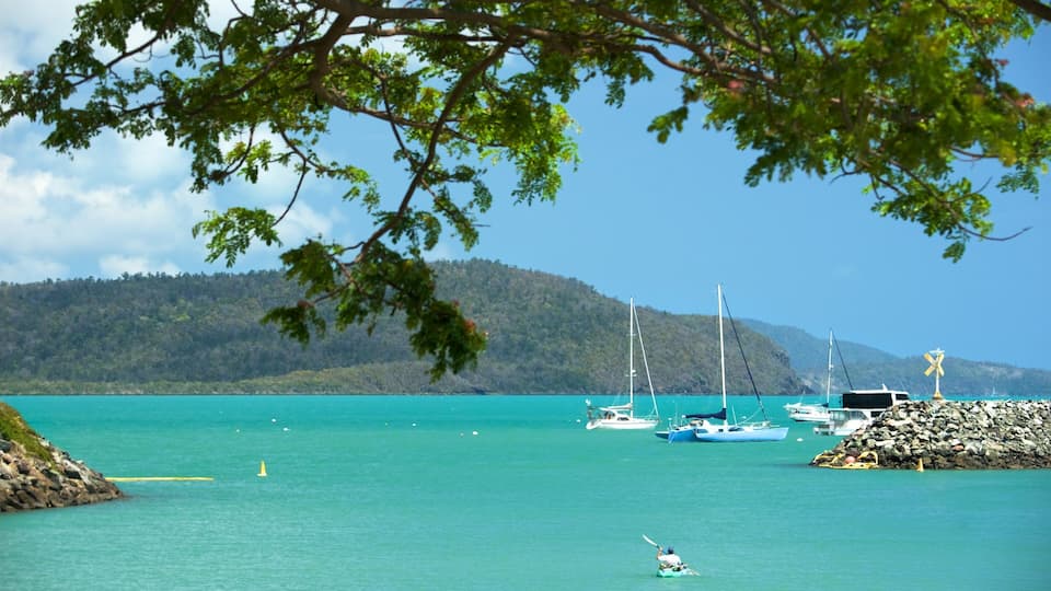 A turquoise sea with boats, a rocky shore, and a forested hill in the background.