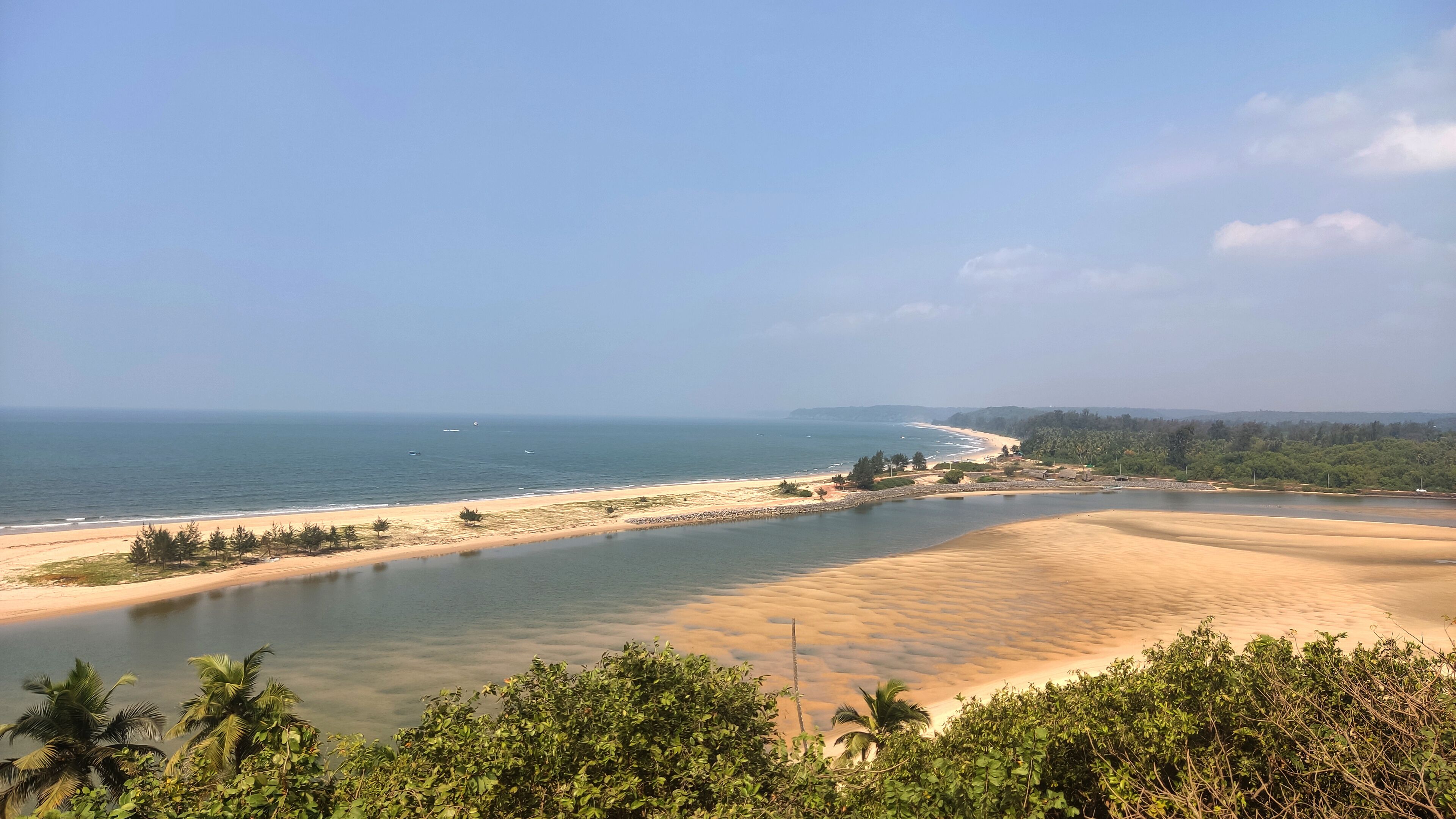 A coastal landscape with a sandy beach, a calm river, and dense greenery.