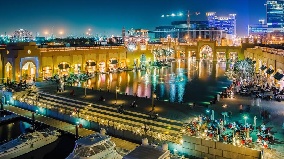 A waterfront promenade with illuminated buildings, a central reflecting pool, and boats docked along the shore.