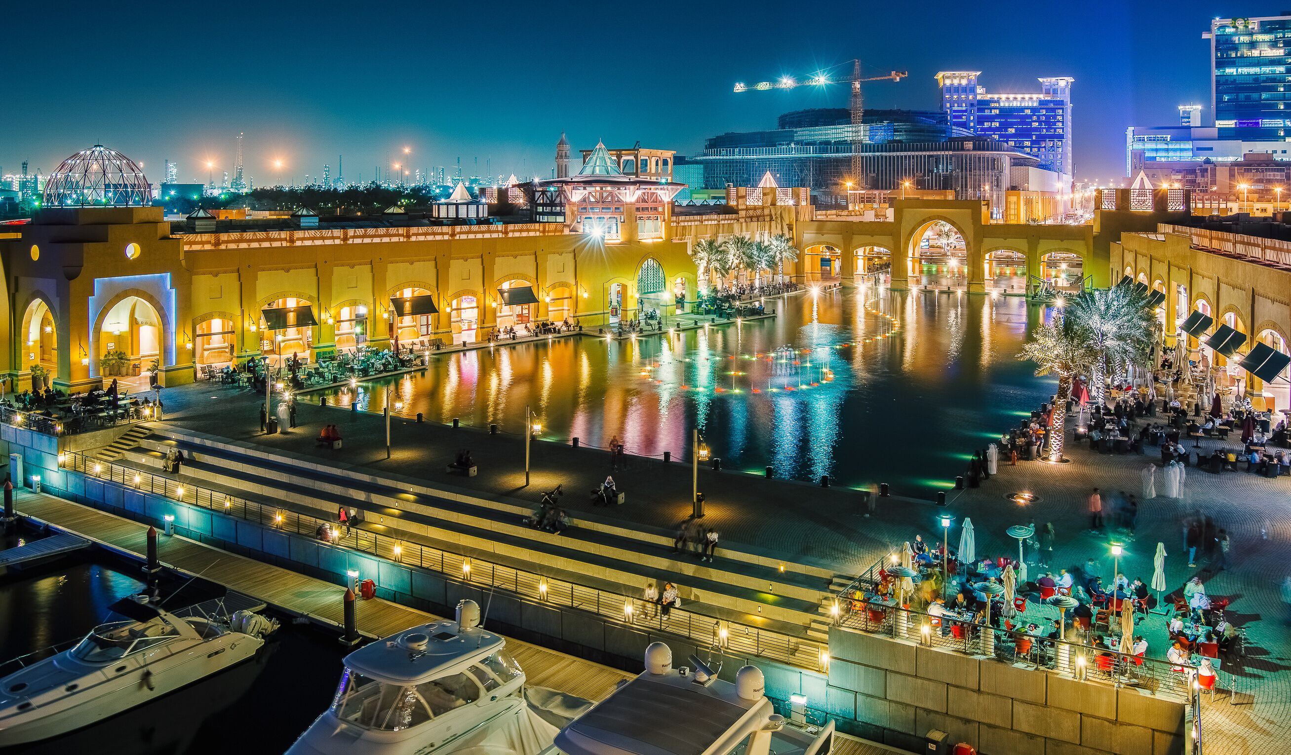A waterfront promenade with illuminated buildings, a central reflecting pool, and boats docked along the shore.