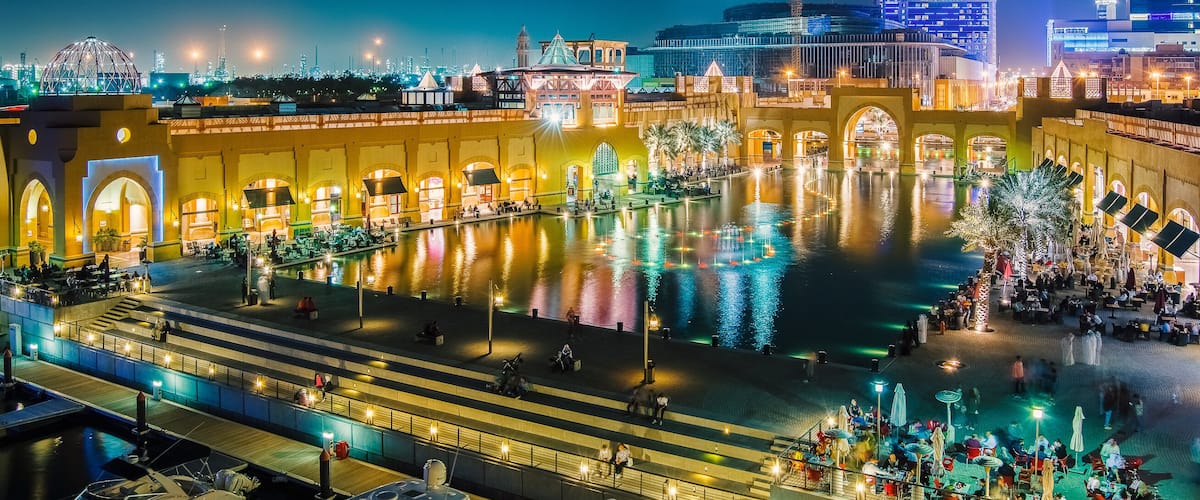 A waterfront promenade with illuminated buildings, a central reflecting pool, and boats docked along the shore.