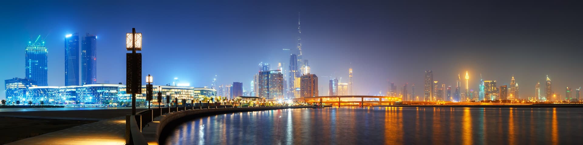 A nighttime cityscape with illuminated buildings and a waterfront promenade.