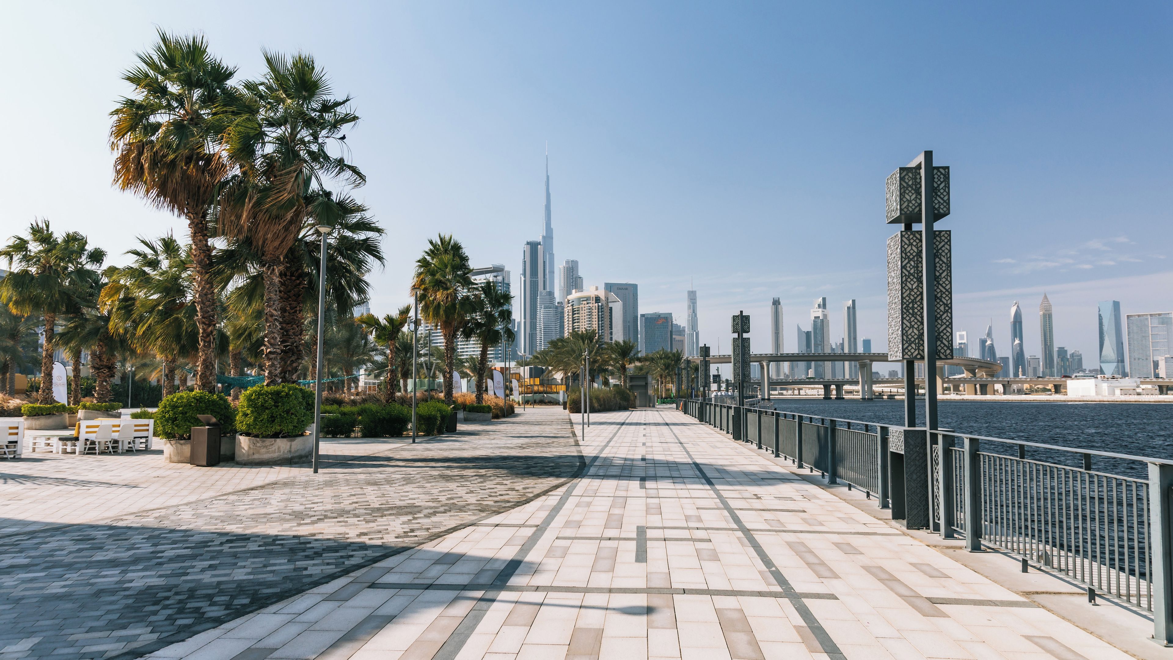 A waterfront promenade with palm trees, a river, and a city skyline including a tall skyscraper.