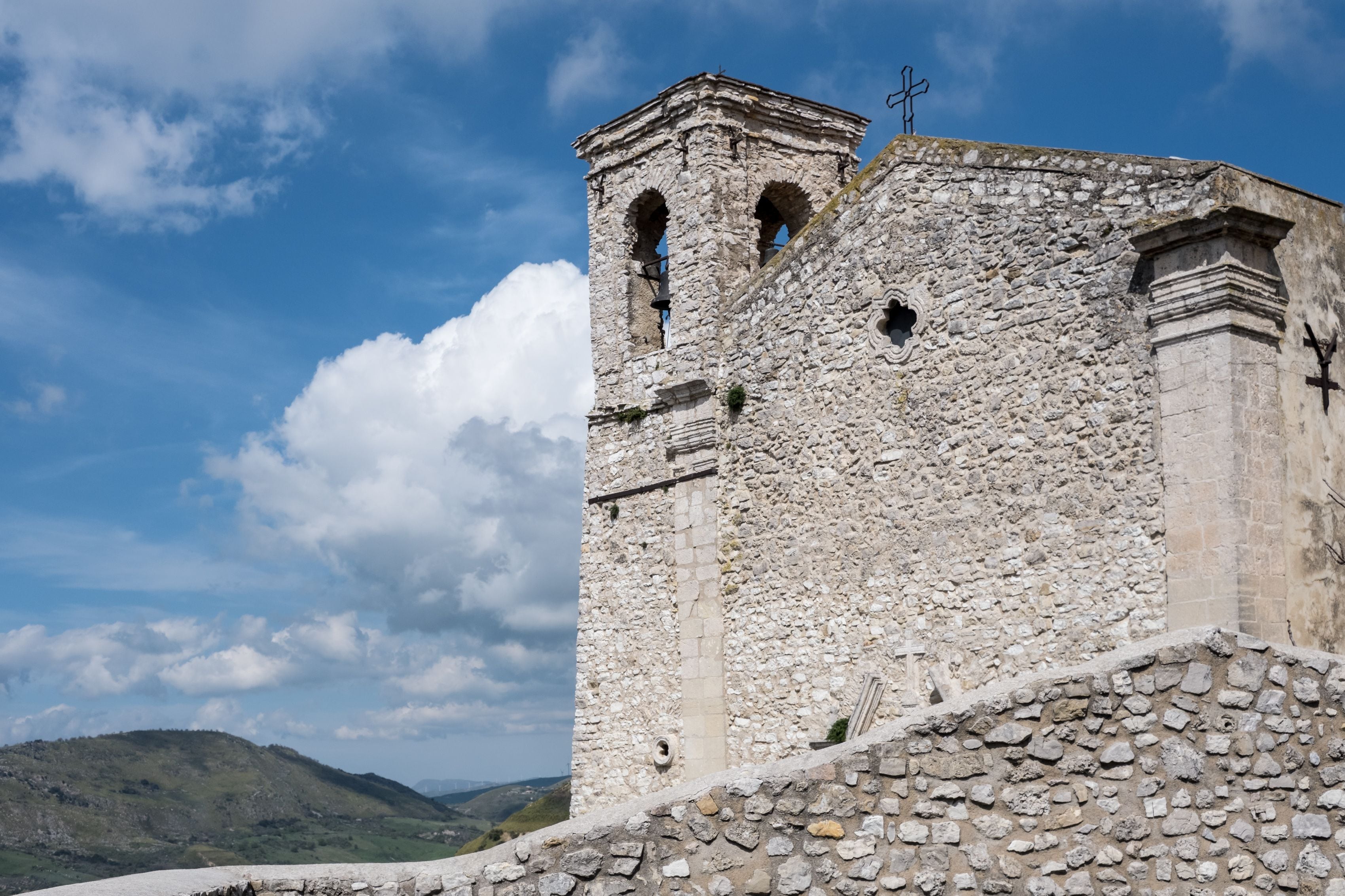 A stone tower with arched openings and a cross on top, set against a clear sky and green hills in the background.