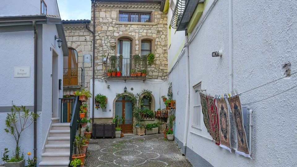 A narrow cobblestone alley with a stone building, potted plants, and hanging laundry.