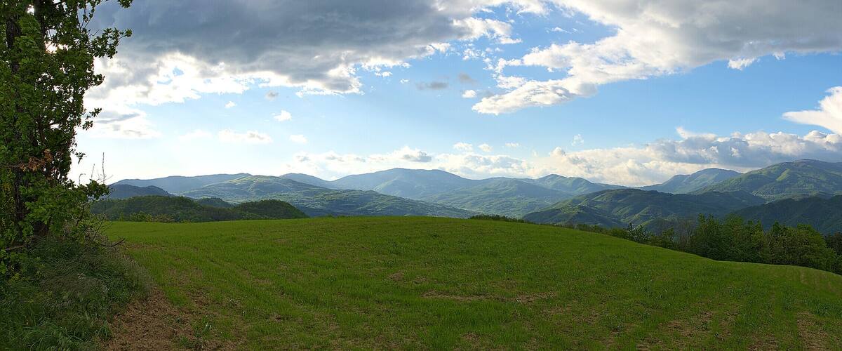 A mountainous landscape with rolling hills, dense forests, and a partly cloudy sky.