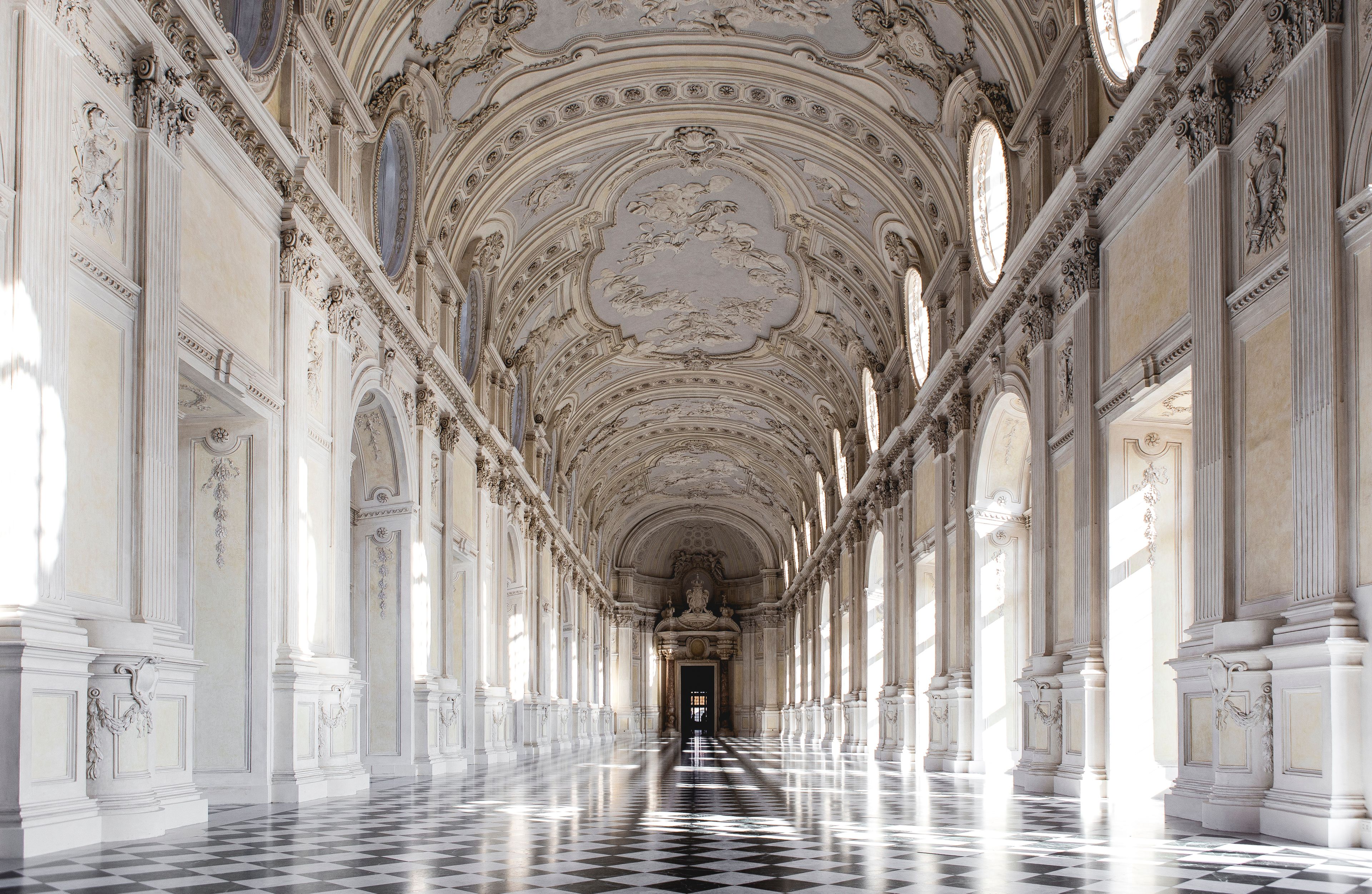 An ornate hallway with intricate ceiling frescoes, classical columns, and a checkered floor.