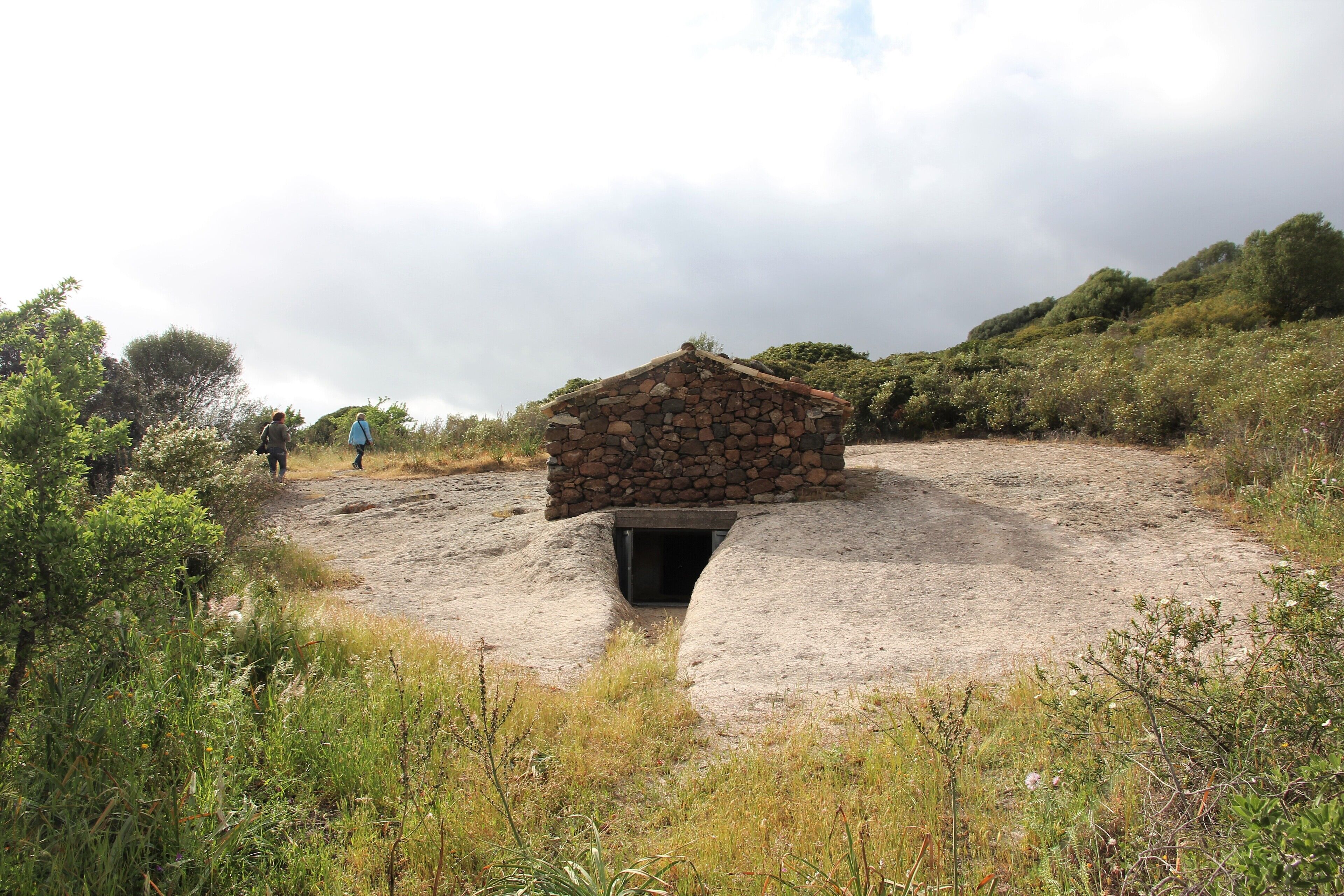 A stone hut with a central opening, surrounded by a natural landscape with vegetation and a cloudy sky.