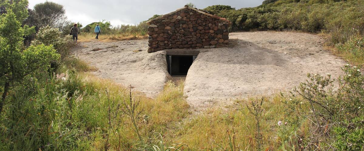 A stone hut with a central opening, surrounded by a natural landscape with vegetation and a cloudy sky.