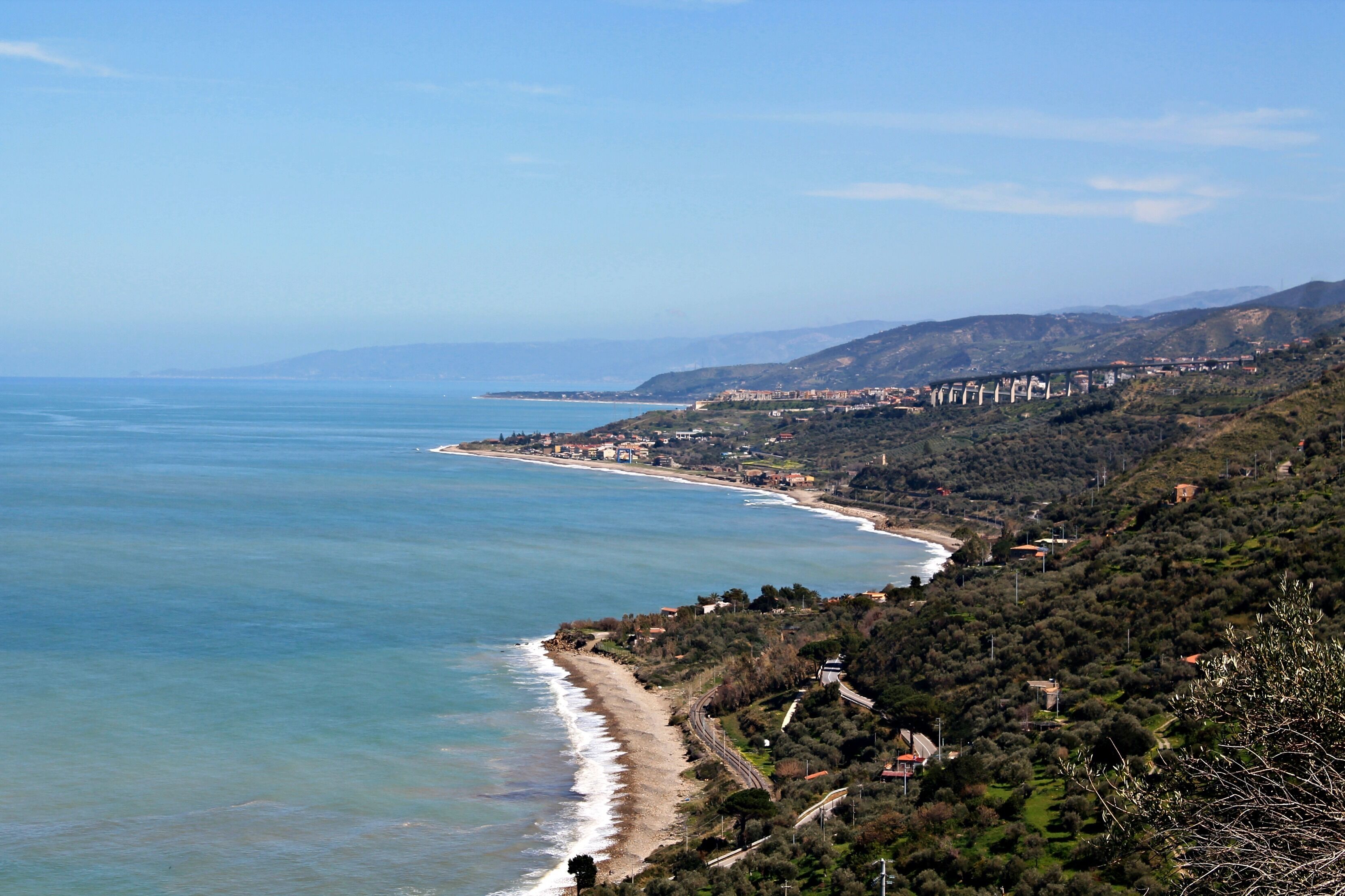 A coastal landscape with a beach, a road, and a hilly area with buildings.