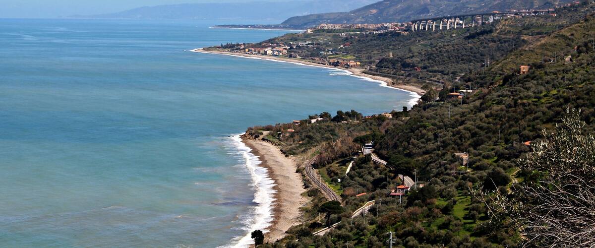 A coastal landscape with a beach, a road, and a hilly area with buildings.