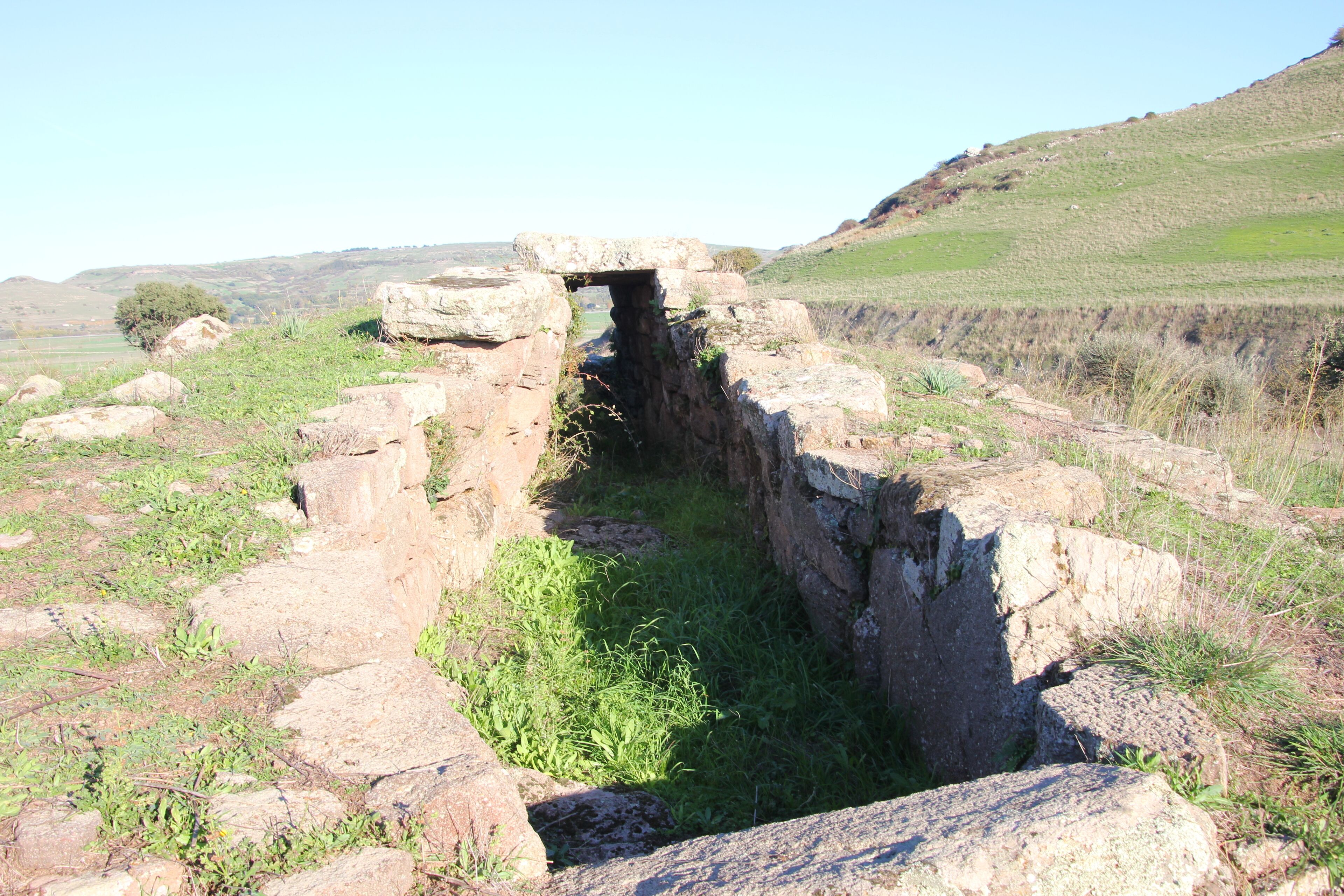 An ancient stone structure with a narrow passageway, surrounded by grassy terrain and large boulders.