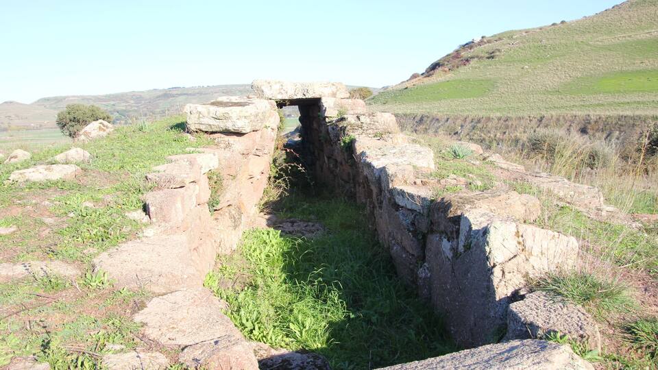 An ancient stone structure with a narrow passageway, surrounded by grassy terrain and large boulders.