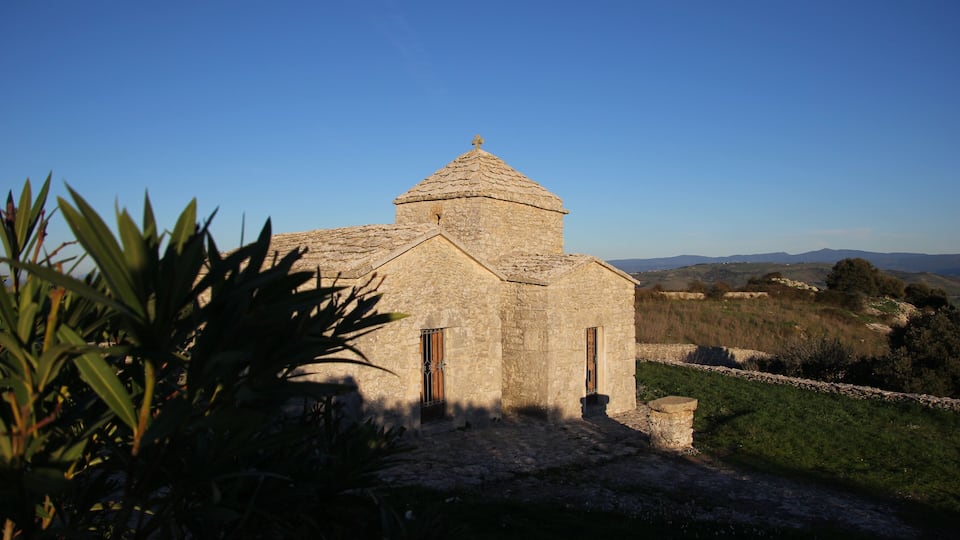 A stone church with a tiled roof and a small bell tower, surrounded by greenery and a clear blue sky.