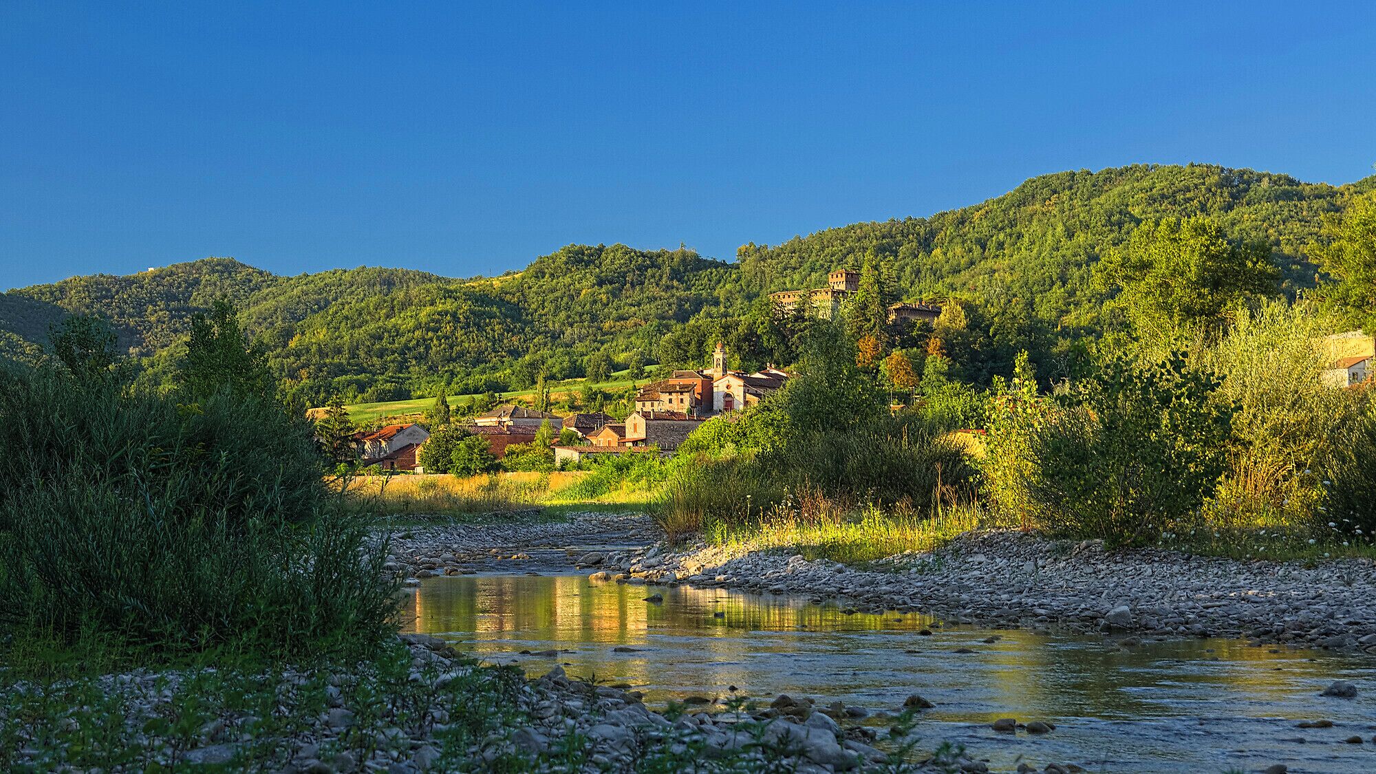 A river with a rocky bed, surrounded by lush greenery and a village with traditional buildings on a hillside.