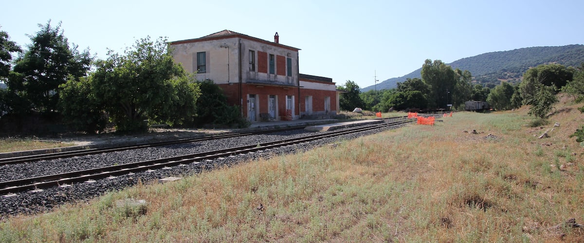 A building with a red roof and white walls, surrounded by trees and a railway track.