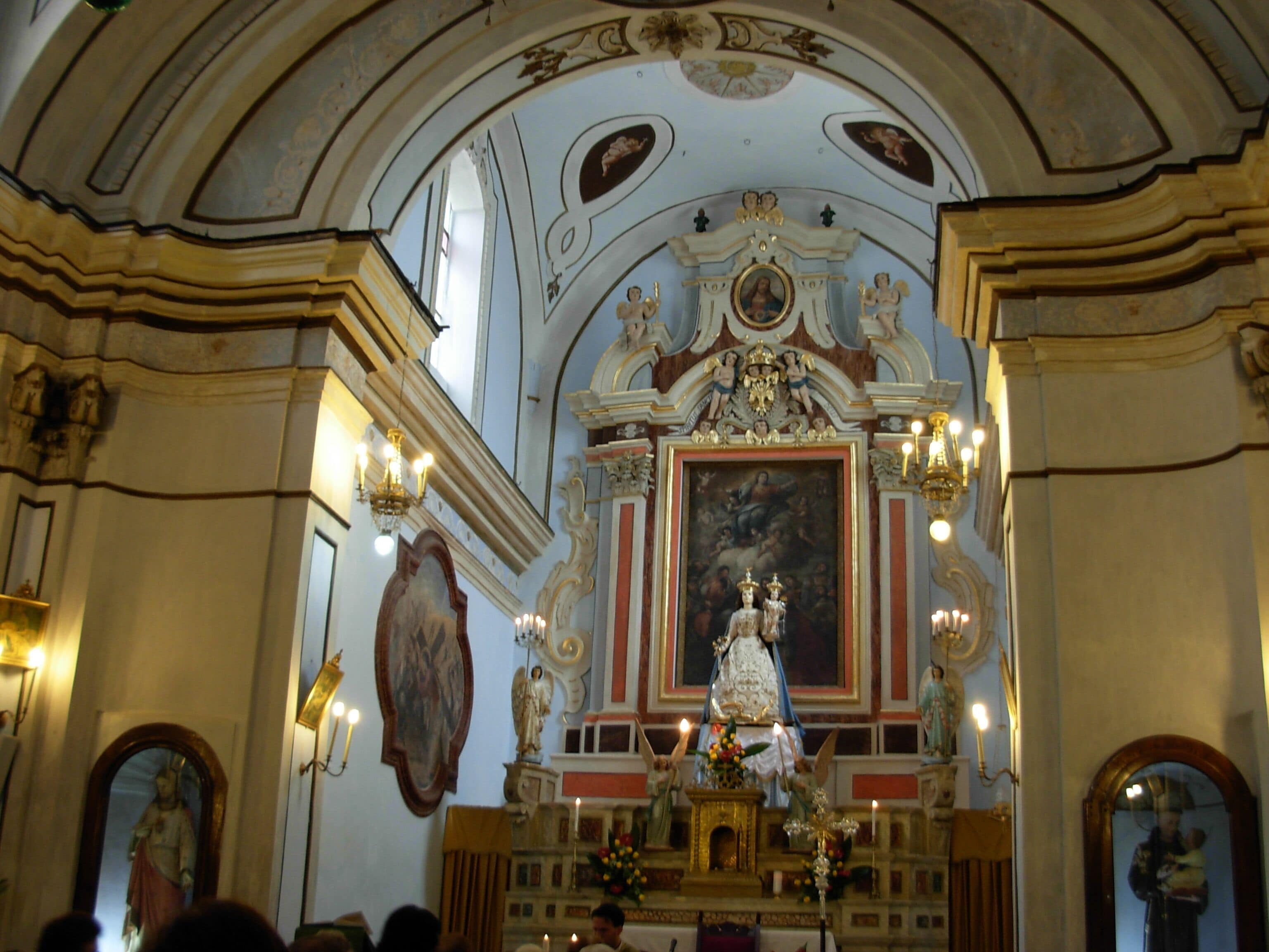 An ornate church interior with a central altar, statues, and a large painting on the wall.