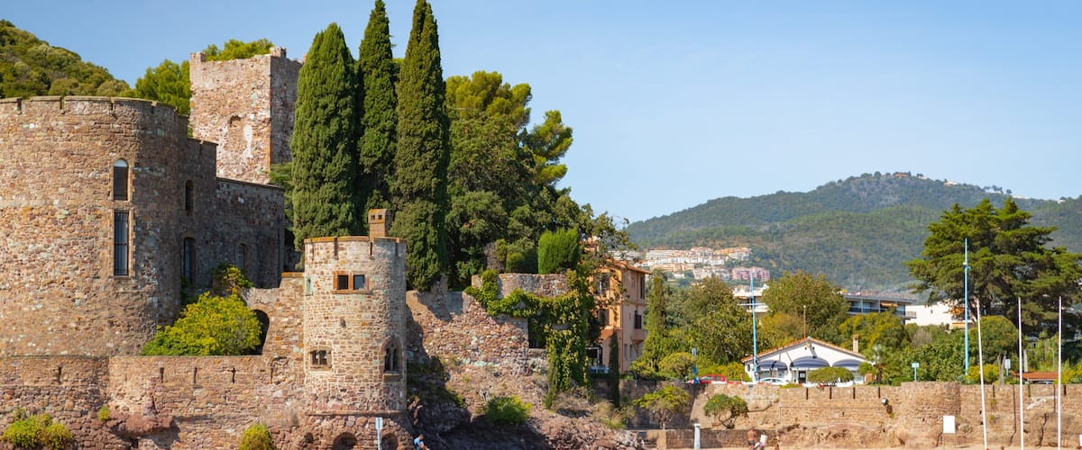 A historic stone castle with towers, a beach, and a hillside town in the background.