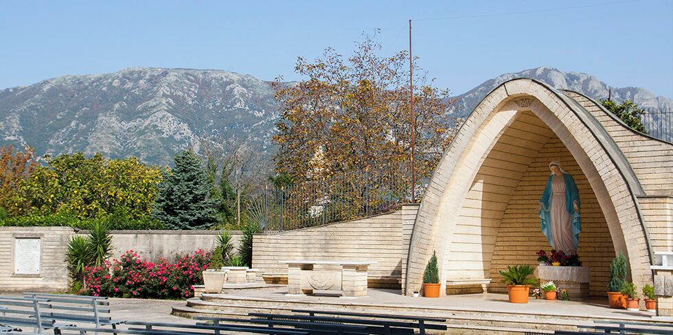 A stone chapel with a statue of a religious figure, surrounded by potted plants and benches, with mountains in the background.
