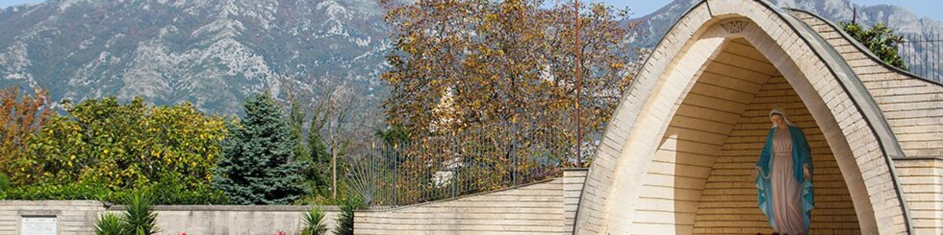A stone chapel with a statue of a religious figure, surrounded by potted plants and benches, with mountains in the background.