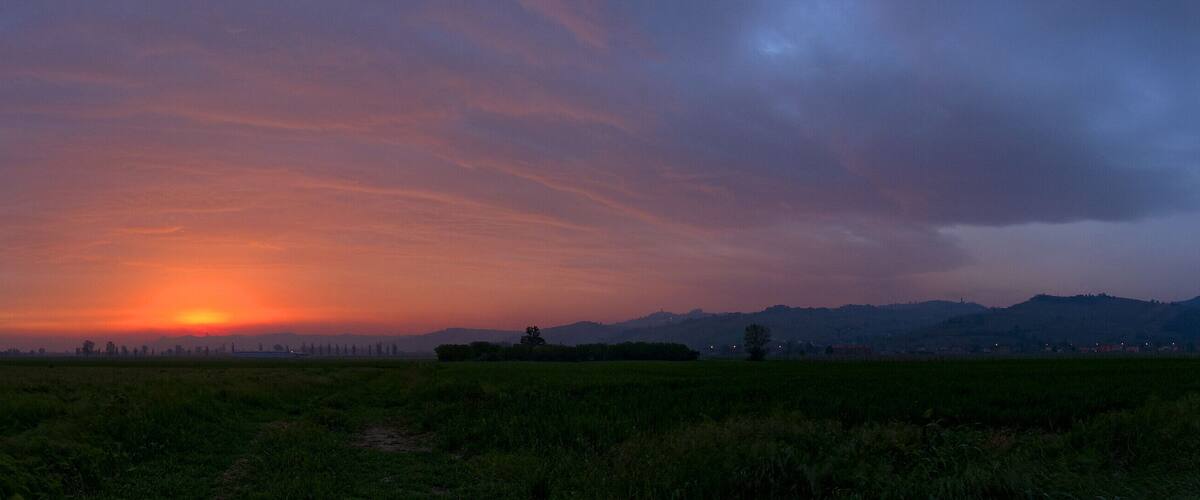 A sunset over a rural landscape with a path leading into the field and distant mountains.
