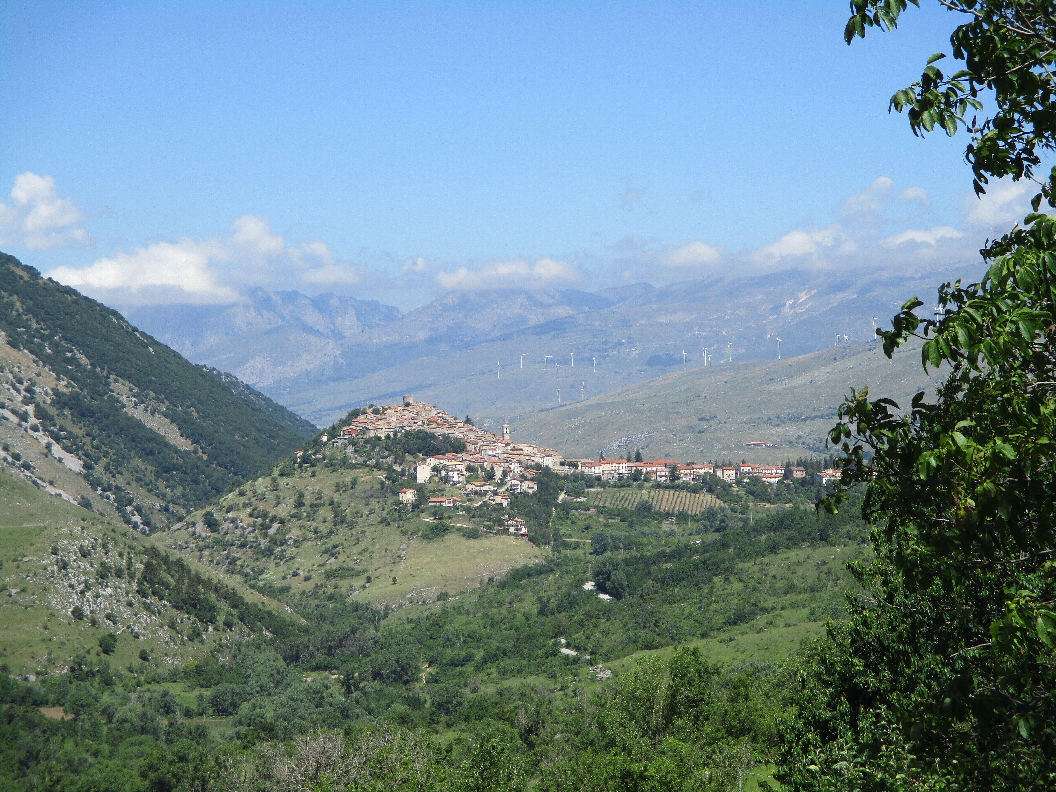 A mountainous landscape with a village, green valleys, and wind turbines in the distance.