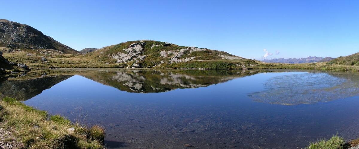 A calm lake surrounded by hills with a clear reflection of the sky and distant mountains.