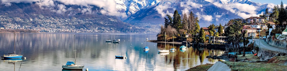 A calm lake with boats, a mountain range with snow-capped peaks, and a small settlement with houses and trees.