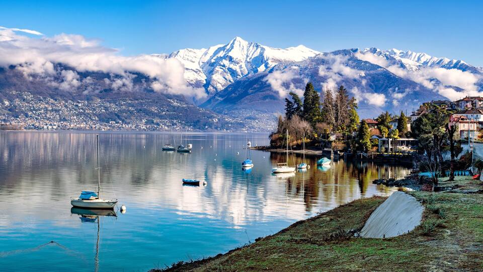 A calm lake with boats, a mountain range with snow-capped peaks, and a small settlement with houses and trees.