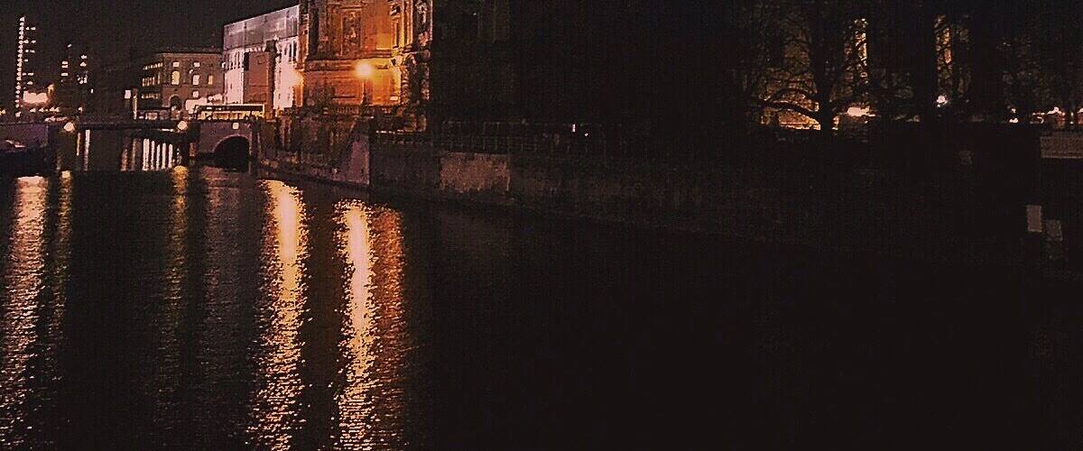 A nighttime view of a historic building with a dome and illuminated windows, reflecting on a body of water.