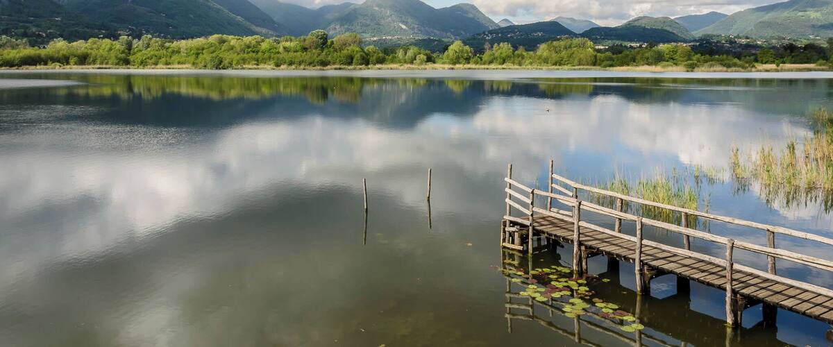 A calm lake with a wooden dock, surrounded by lush green hills and mountains under a partly cloudy sky.
