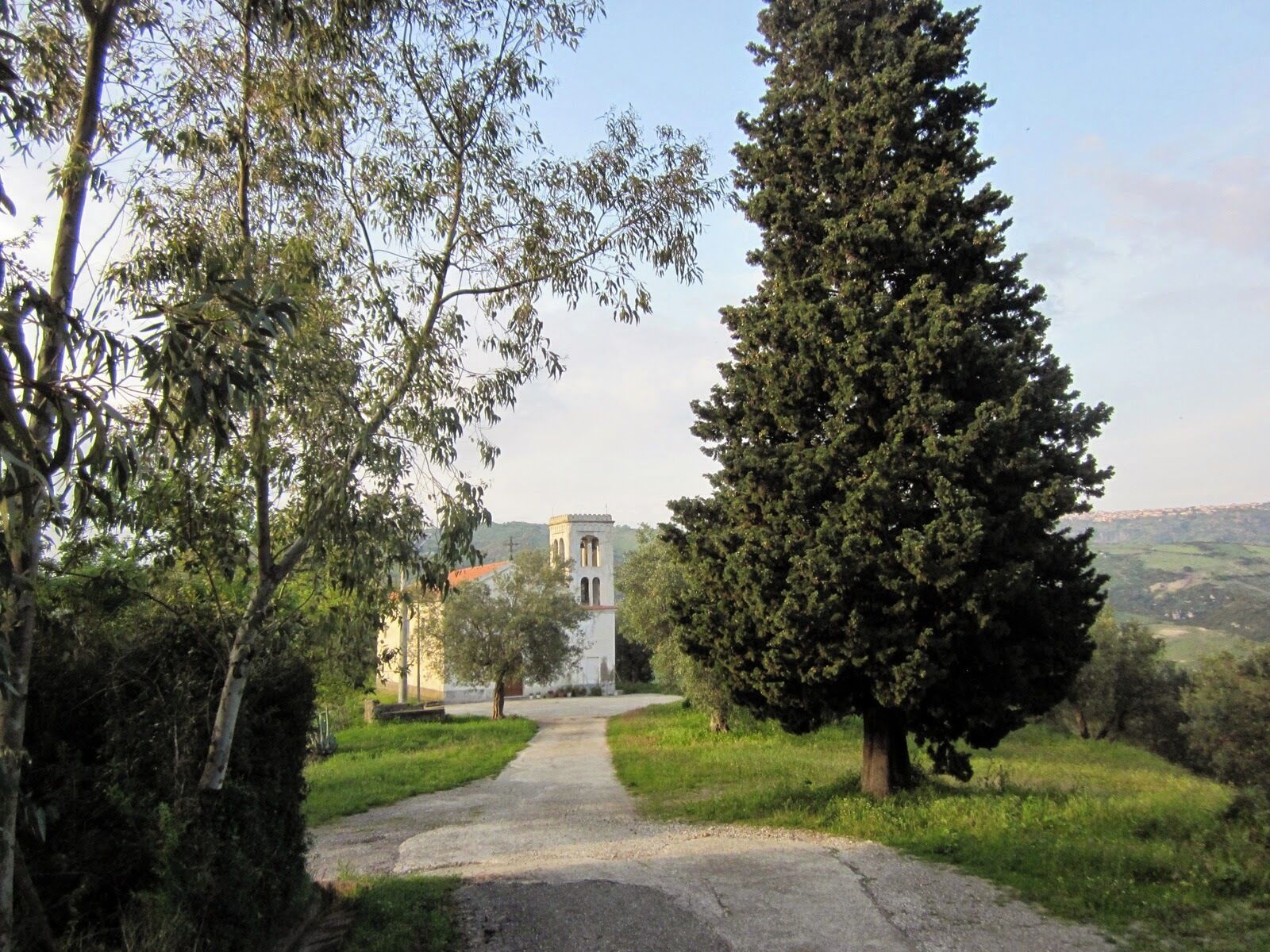 A pathway leading to a white church with a bell tower, surrounded by trees and greenery.