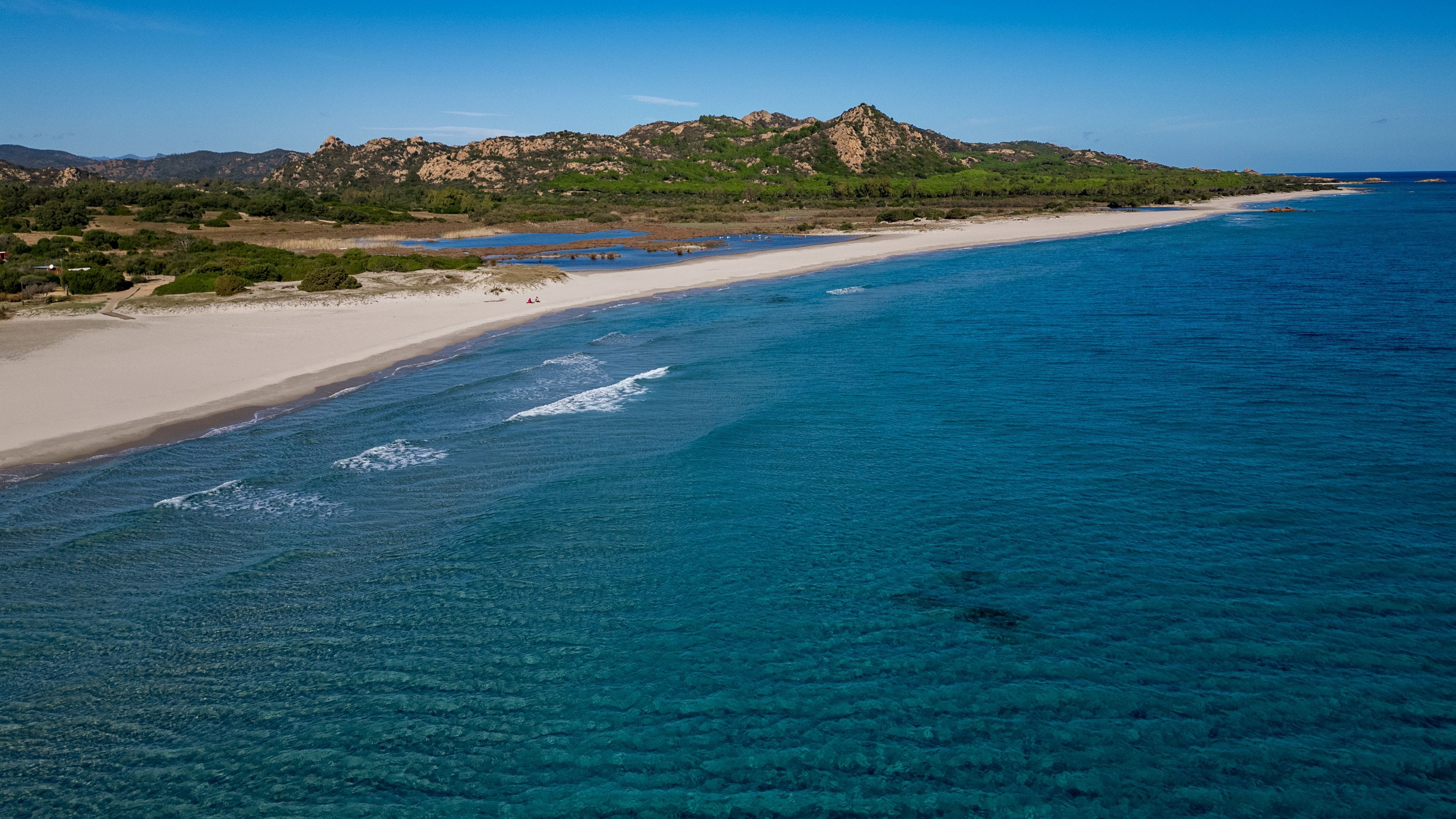 Ett kustlandskap med ett klart, blått hav, en sandstrand och ett bergskedja i bakgrunden.