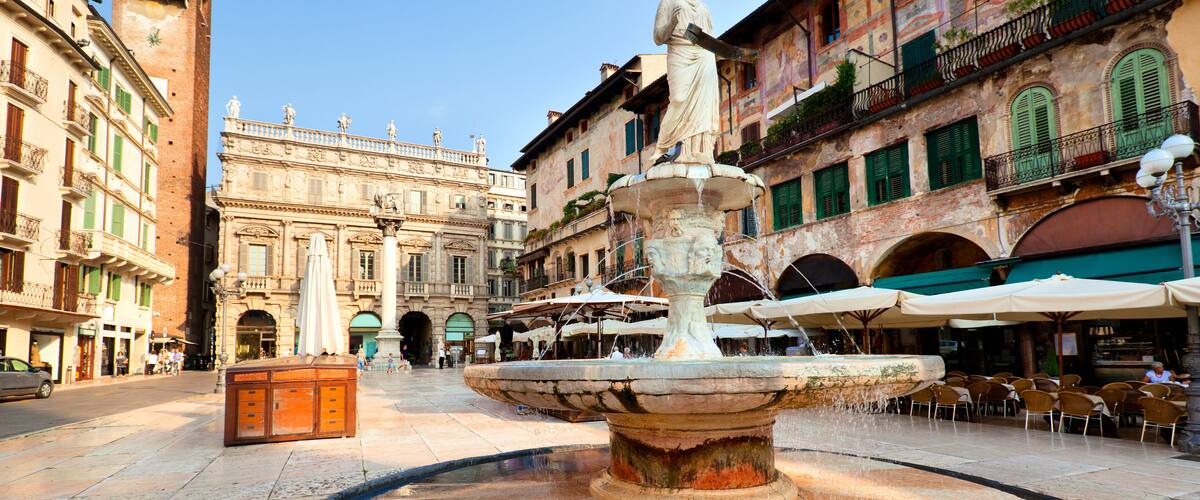 A historic fountain with a statue in the center, surrounded by classic European architecture and outdoor seating areas.