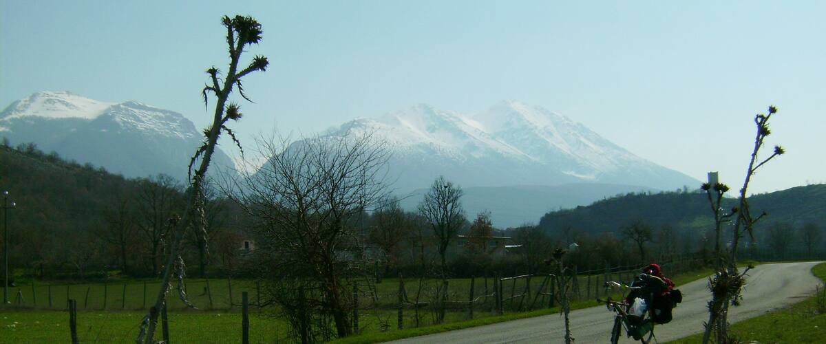 A rural road with a cyclist, snow-capped mountains in the background, and sparse vegetation.