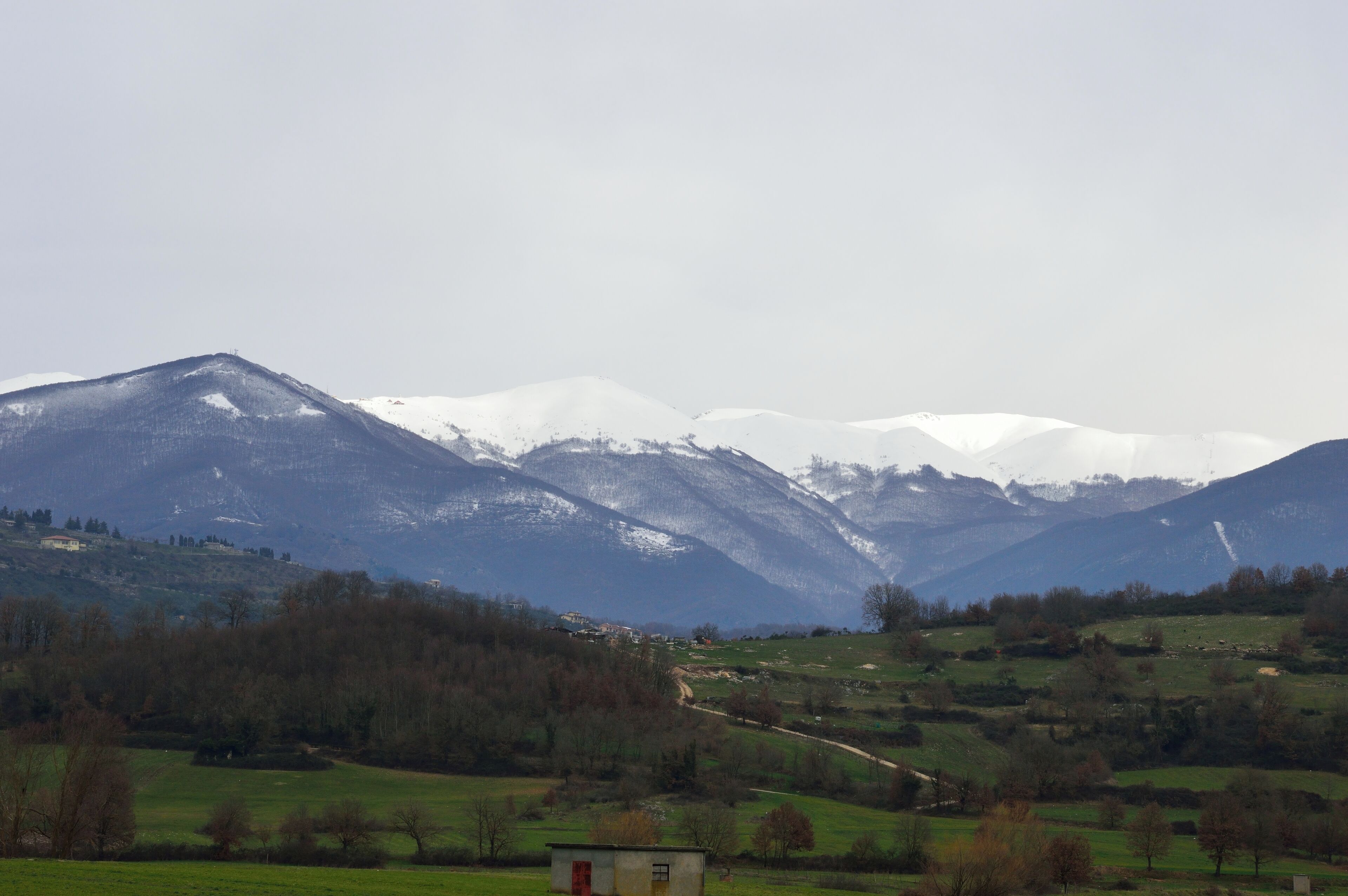 A mountainous landscape with snow-capped peaks, a valley with scattered trees, and a small building in the foreground.