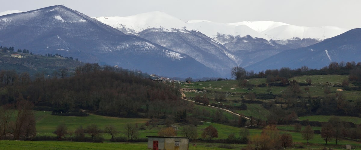 A mountainous landscape with snow-capped peaks, a valley with scattered trees, and a small building in the foreground.