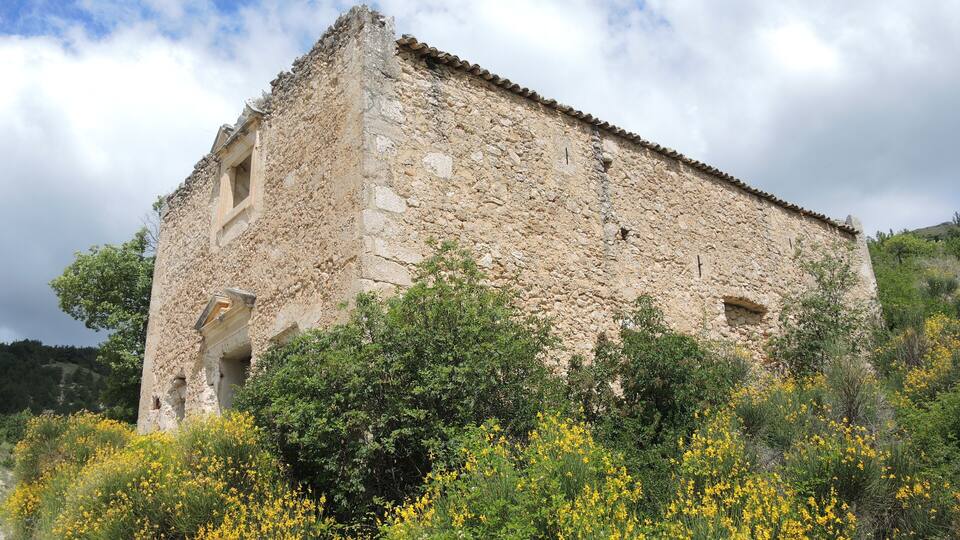 A stone building with a tiled roof surrounded by dense greenery and yellow flowers.