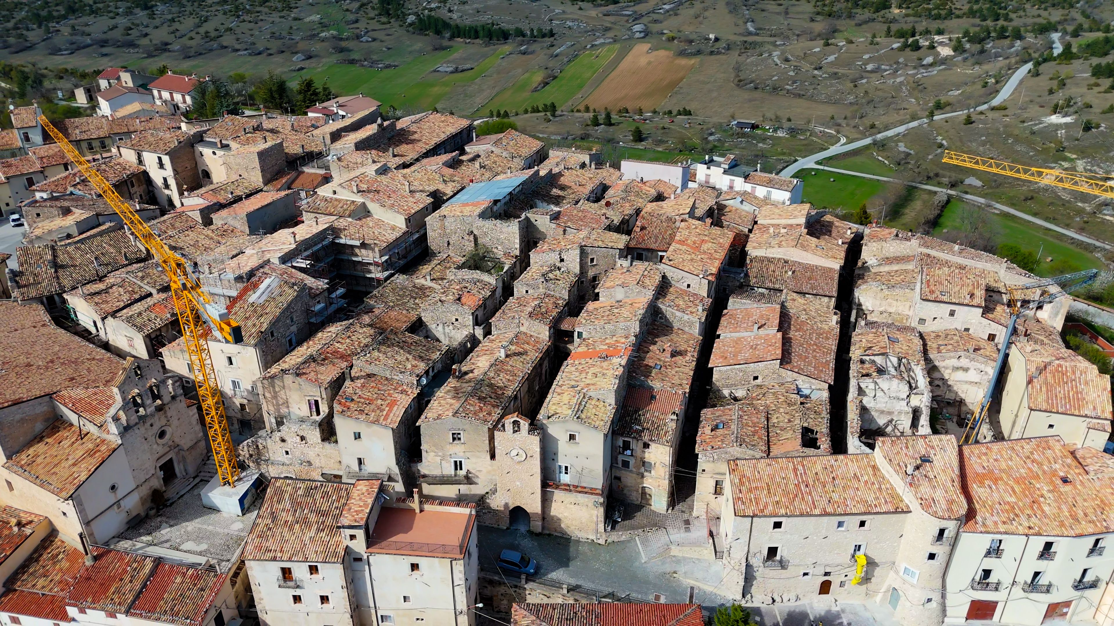 An aerial view of a historic town with terracotta-roofed buildings, a construction crane, and a winding river in the background.