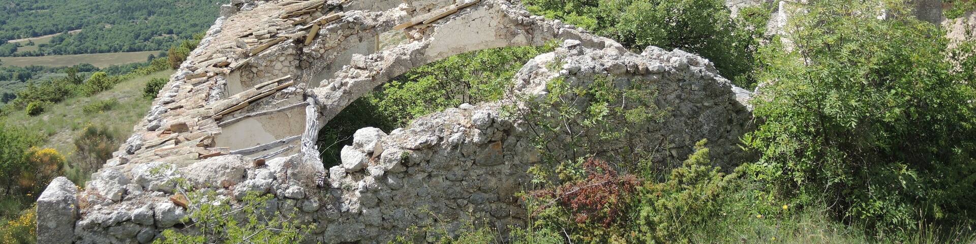 An ancient stone structure with a partially collapsed roof, surrounded by lush green hills and a clear blue sky.
