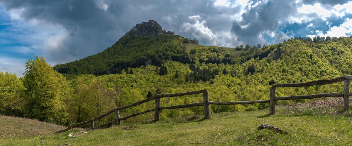 A grassy field with a wooden fence, a forested hill, and a prominent rocky peak under a cloudy sky.