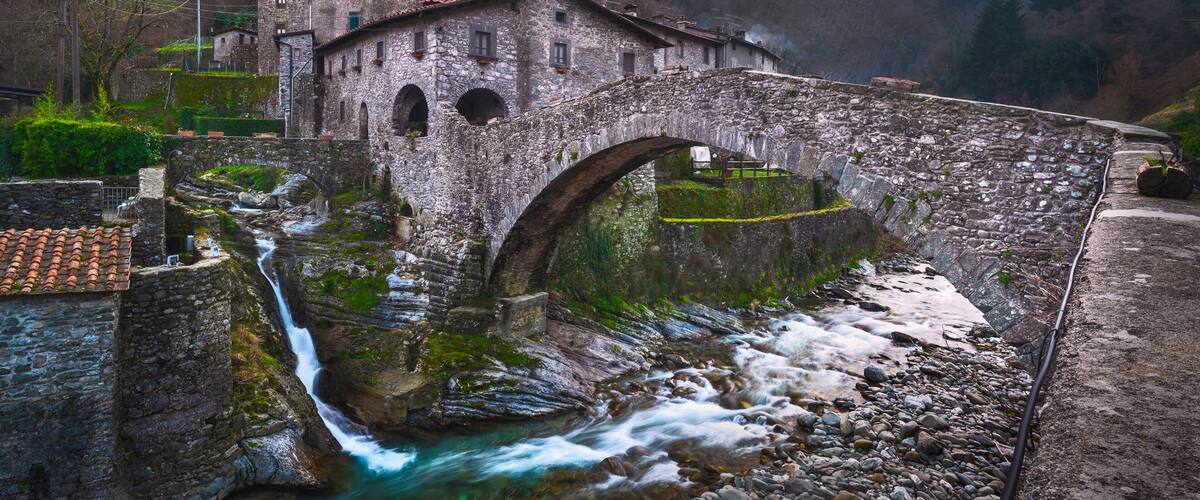 A stone bridge over a river, surrounded by stone buildings and a mountainous backdrop.