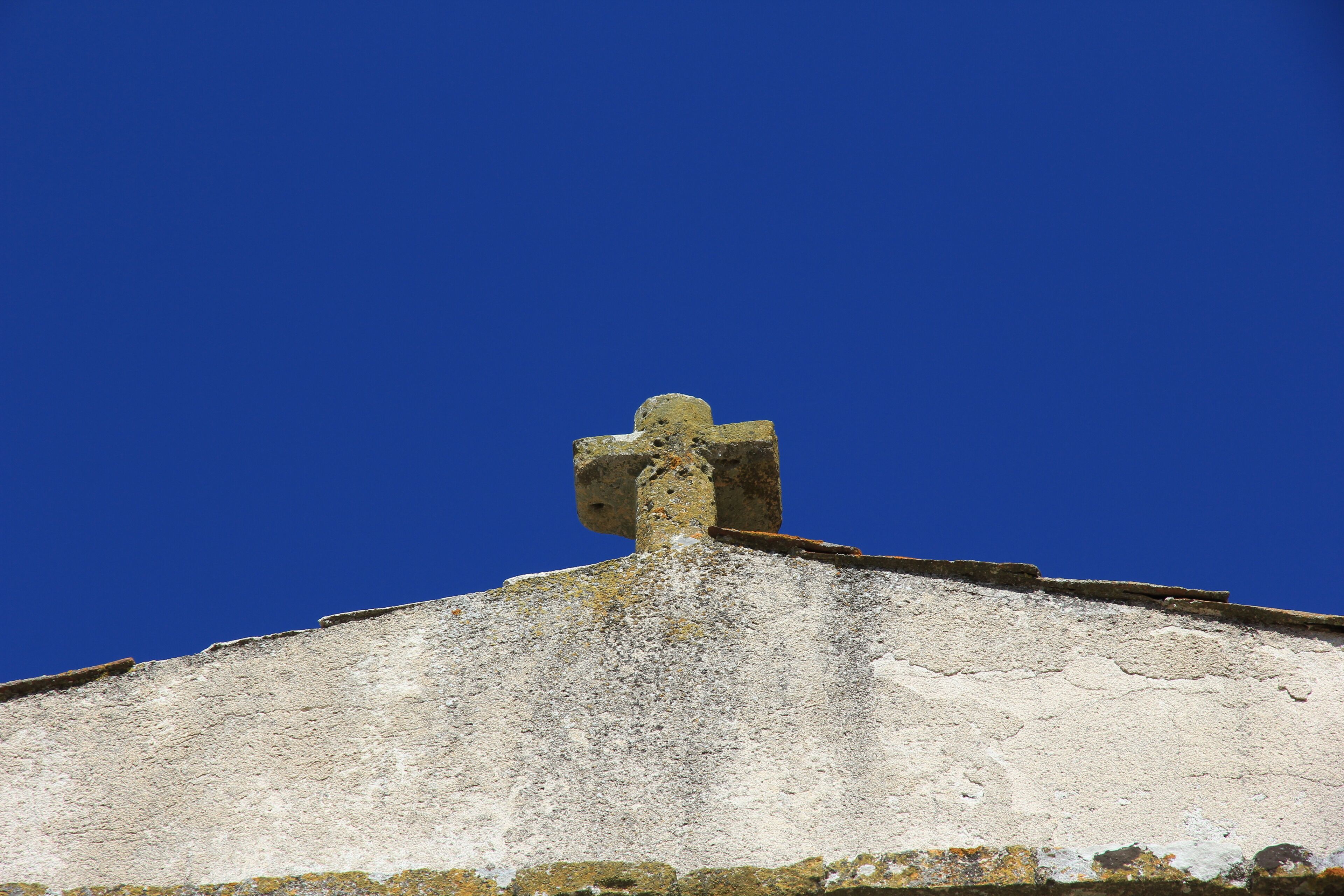 A weathered stone cross atop a building with a clear blue sky in the background.