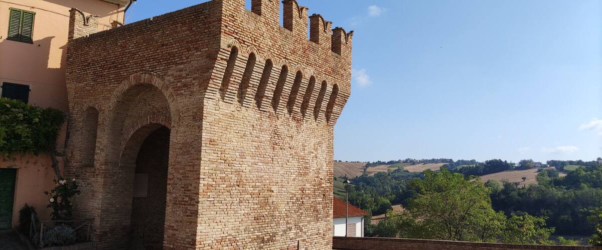 A historic brick tower with arched openings and battlements, set against a backdrop of rolling hills and a clear sky.