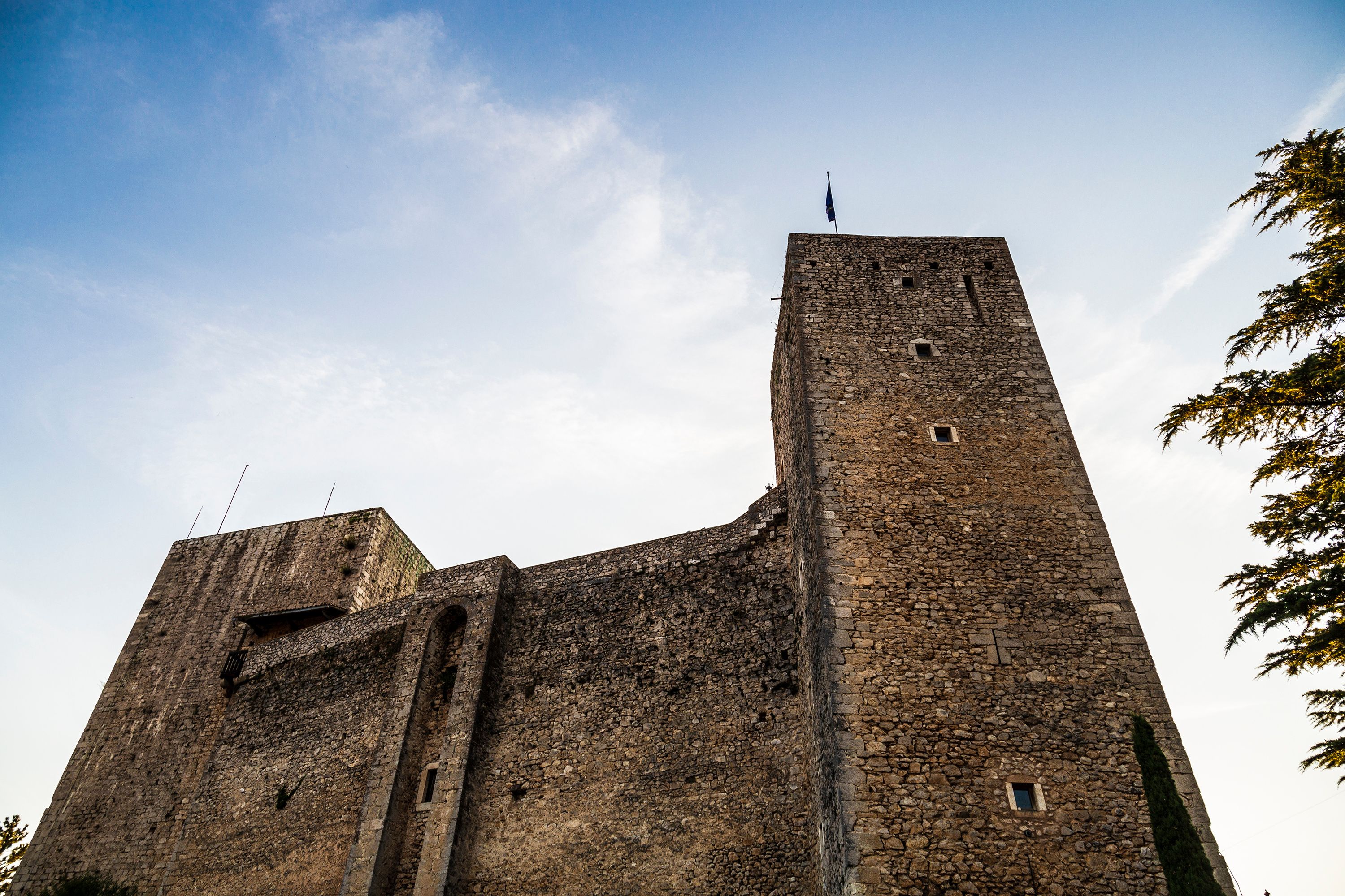 A tall, historic stone tower with a pointed roof, surrounded by clear blue sky and sparse clouds.