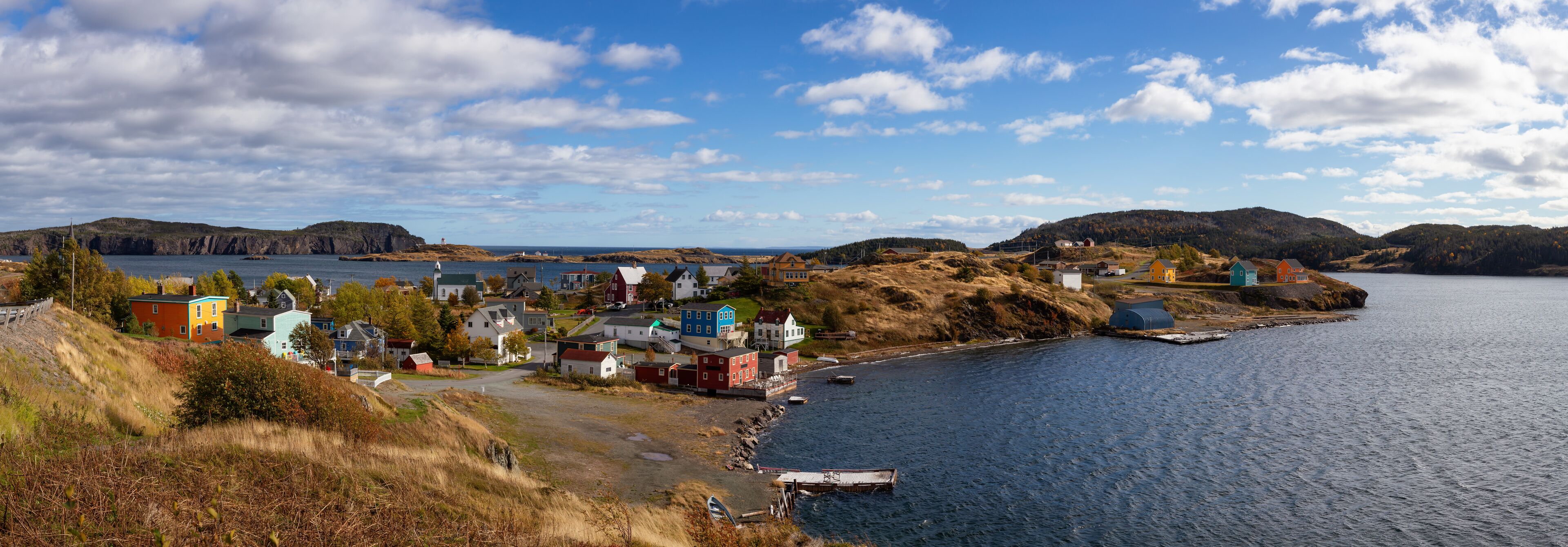 A coastal town with colorful houses by the water, a lighthouse, and hills in the background.