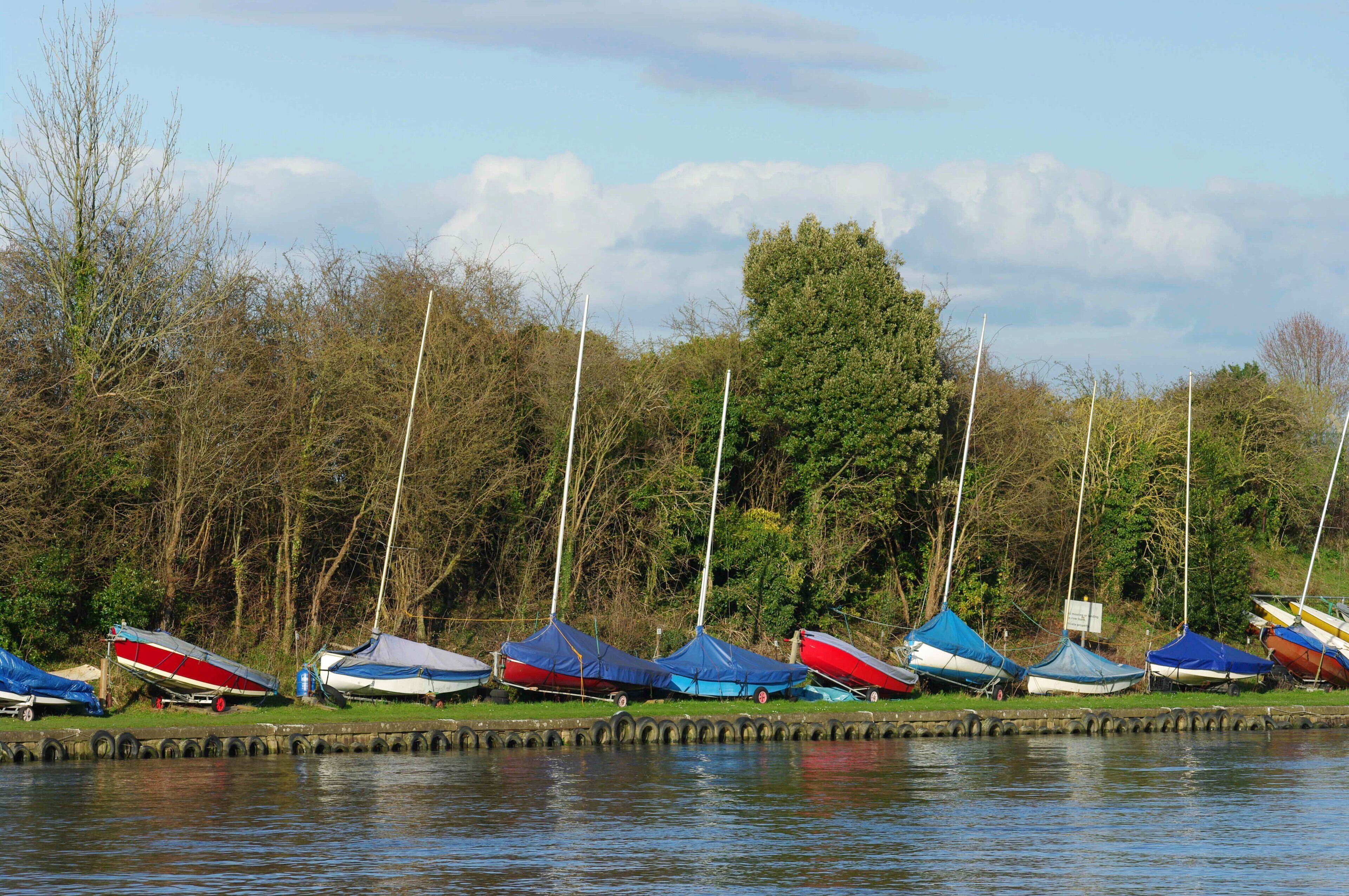 A row of boats covered with tarps, docked along a calm water body with trees and a cloudy sky in the background.