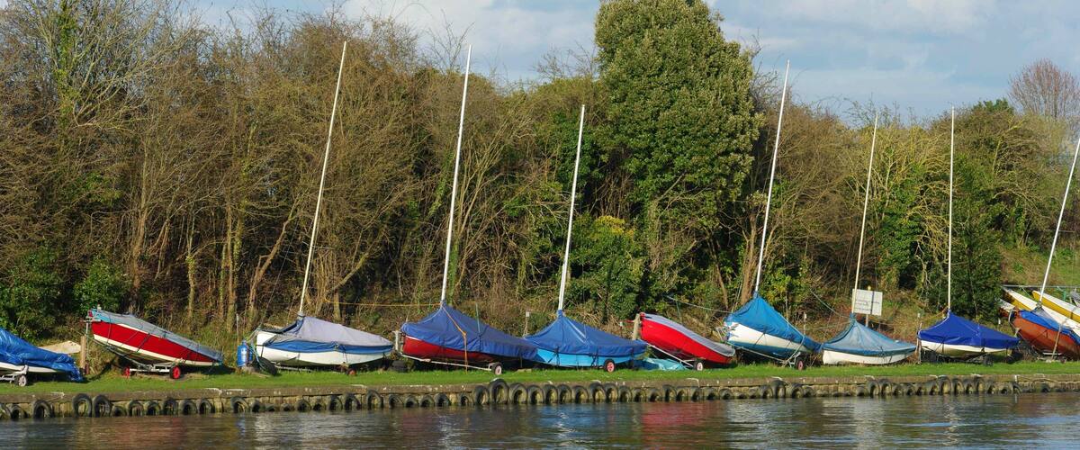 A row of boats covered with tarps, docked along a calm water body with trees and a cloudy sky in the background.