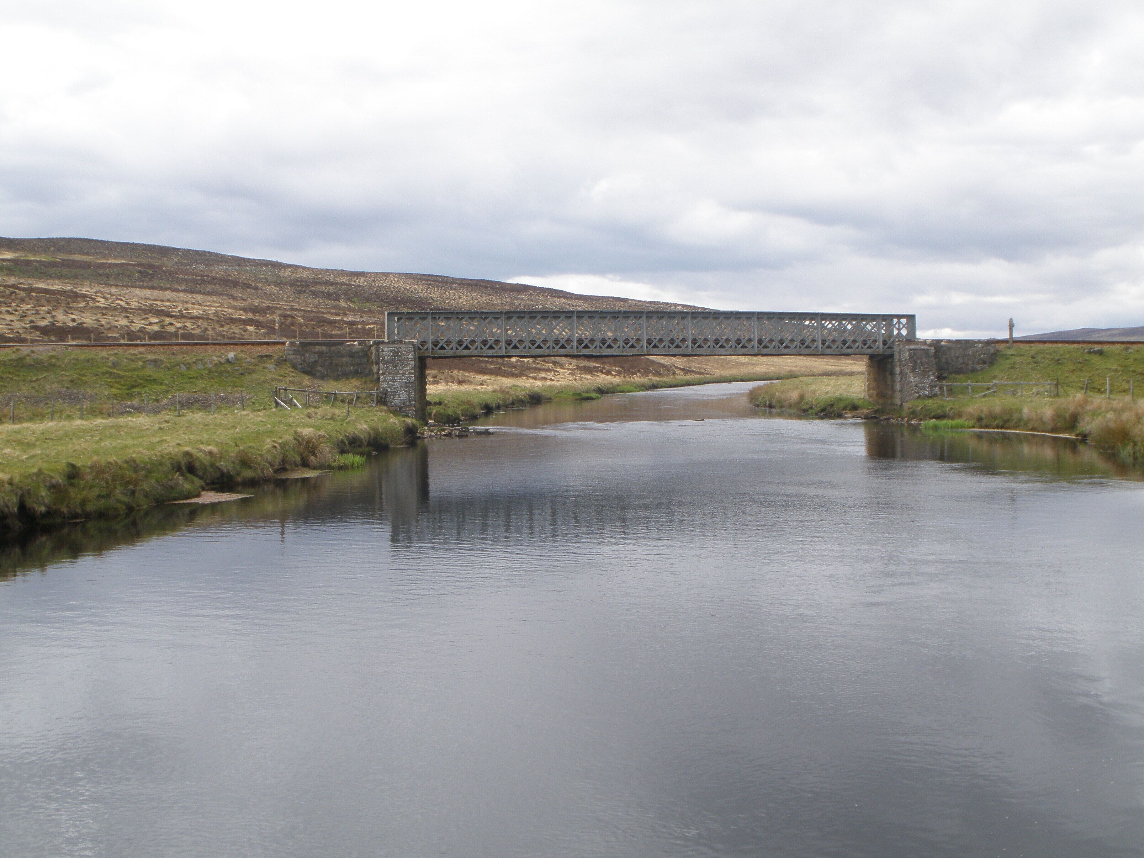 A metal bridge over a calm river with grassy banks and hills in the background.