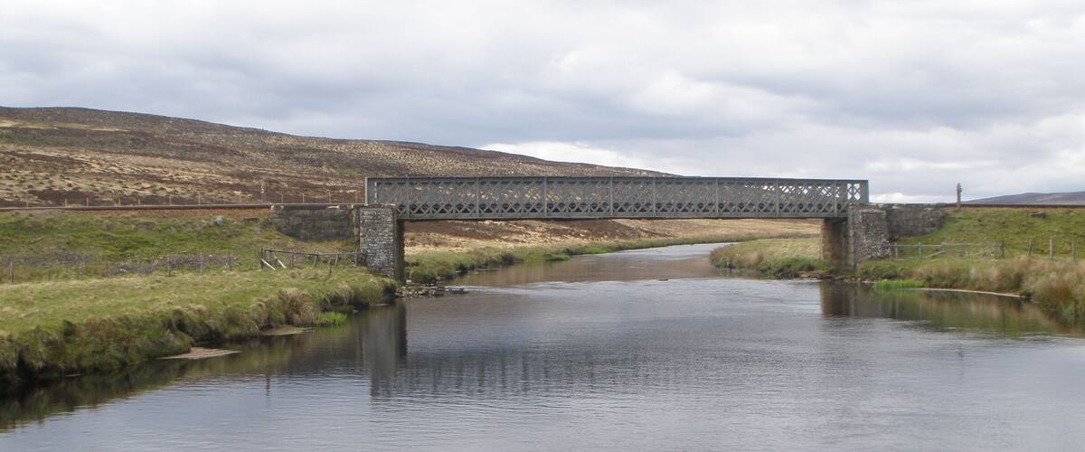 A metal bridge over a calm river with grassy banks and hills in the background.